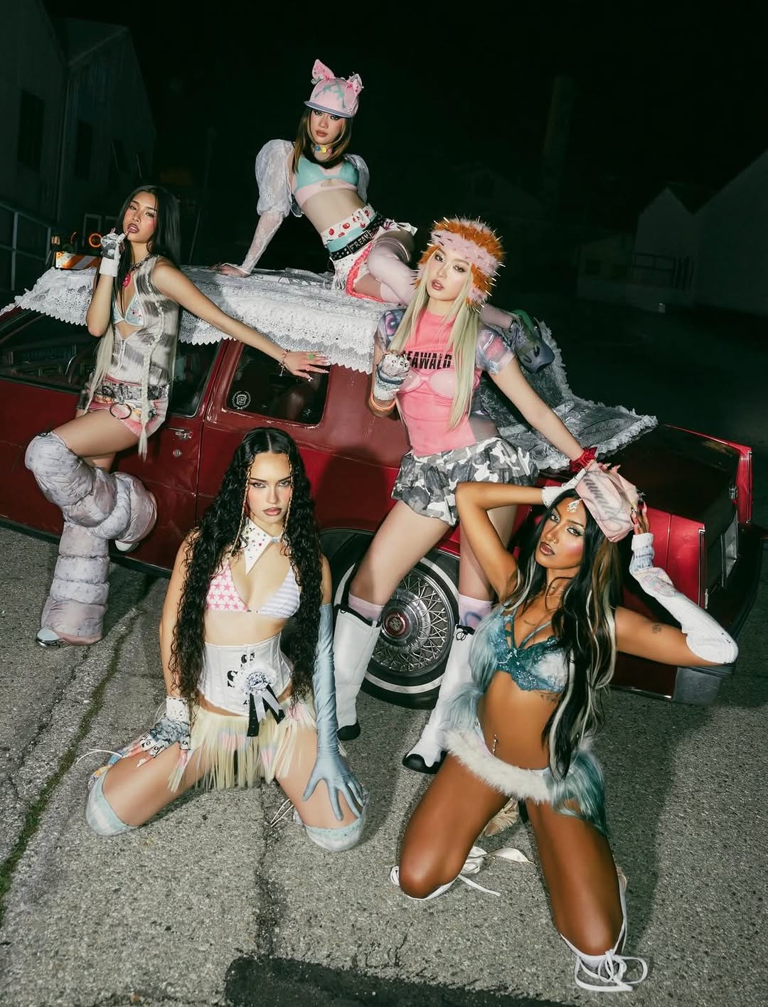 Girl group Katseye posed in colorful outfits in front of a red car outside at night